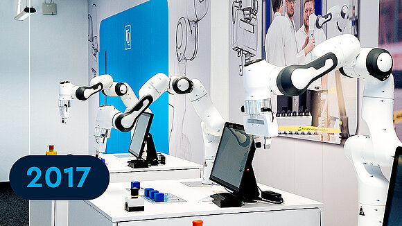 Three robotic arms on tables and pictures in the background showing the year 2017