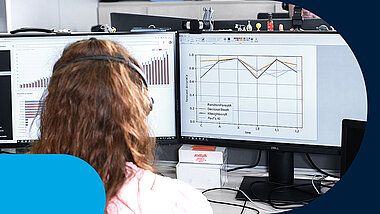 A female employee seen from behind, standing in front of a desk with two monitors