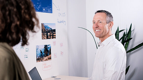 A man is standing in front of a wall, looking toward another desk in the office