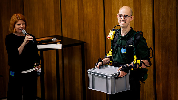 Peter Schaefer mit EASE Exoskelett und Kiste bei Bühnenvortrag