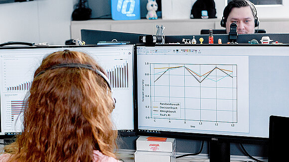 A female employee seen from behind, standing in front of a desk with two monitors