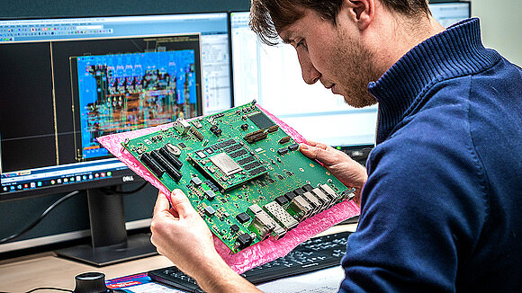 A man holds a circuit board in his hands in front of a PC screen