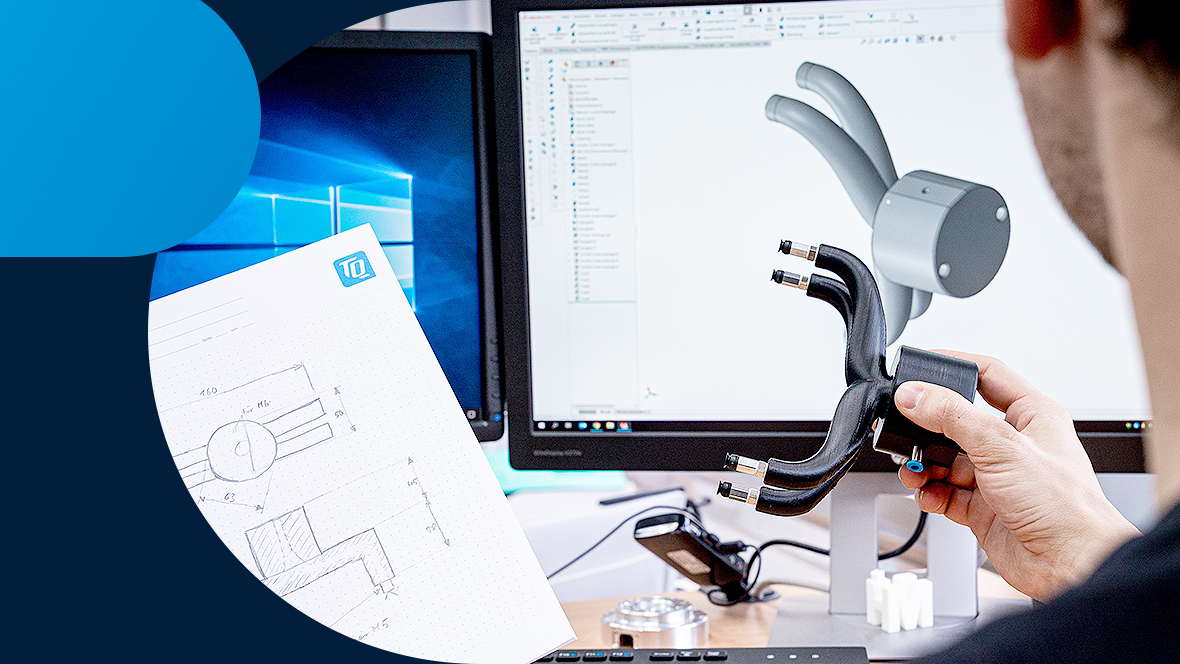 A person looks at a component and a drawing in front of a PC screen