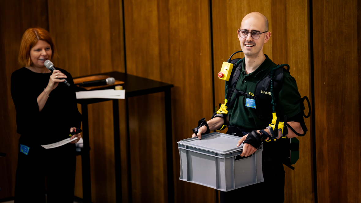 Peter Schaefer mit EASE Exoskelett und Kiste bei Bühnenvortrag